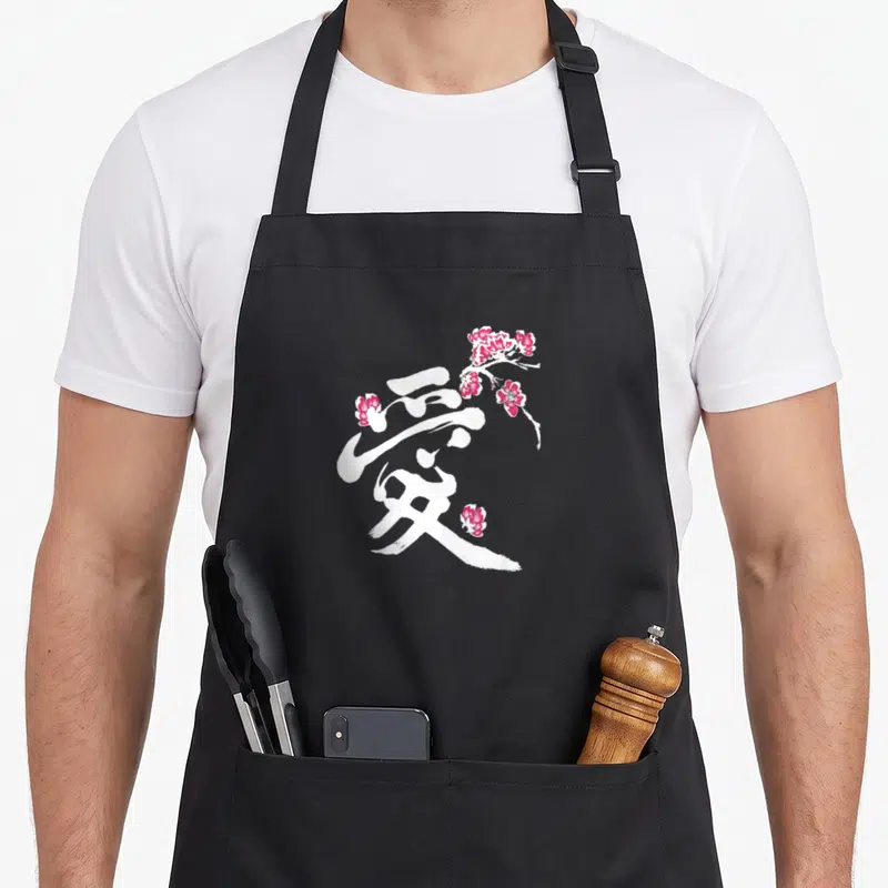 Tablier de cuisine noir avec le Kanji Amour blanc entouré de fleurs de cerisier Sakura roses stylisées.