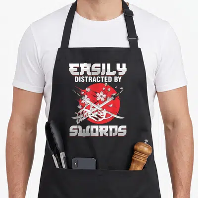 Tablier de cuisine noir avec deux katanas croisés, masque blanc et fleurs de sakura sur cercle rouge.