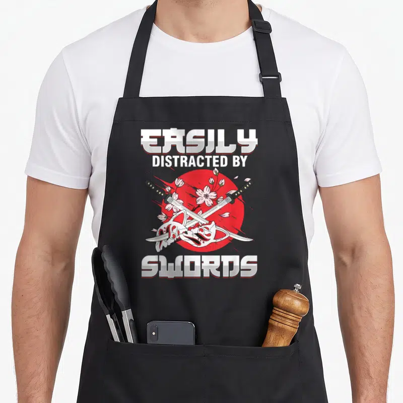 Tablier de cuisine noir avec deux katanas croisés, masque blanc et fleurs de sakura sur cercle rouge.