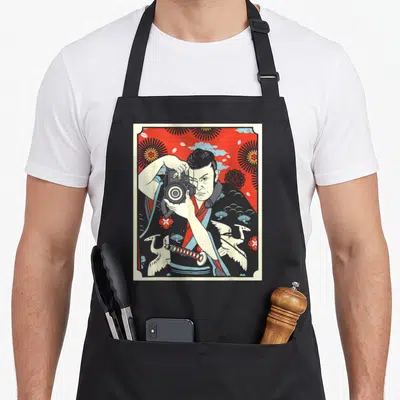 Tablier de cuisine noir avec illustration d'un photographe samouraï japonais tenant une caméra sur fond rouge et motifs floraux traditionnels.