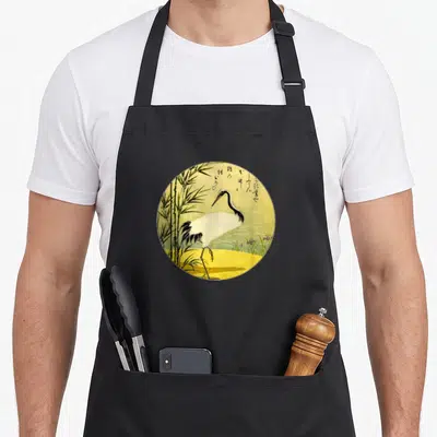 Tablier de cuisine noir avec une illustration circulaire représentant une grue blanche au milieu de tiges de bambou vertes sur fond jaune.