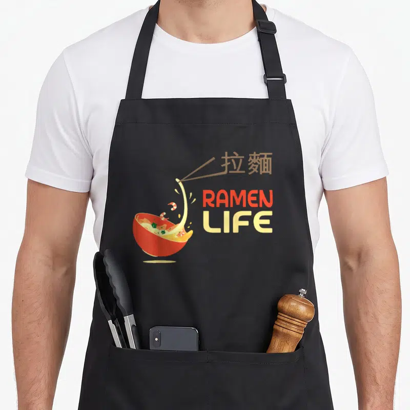 Tablier de cuisine noir avec illustration d'un bol de ramen rouge, chopsticks et texte RAMEN LIFE coloré.
