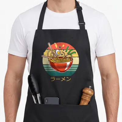 Tablier de cuisine noir avec illustration d'un bol de ramen aux noodles, œuf et baguettes sur fond circulaire coloré.