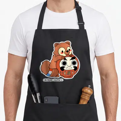 Tablier de cuisine noir avec illustration d'un Tanuki marron posé sur une poupée Daruma rouge et blanche.