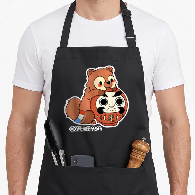 Tablier de cuisine noir avec illustration d'un Tanuki marron posé sur une poupée Daruma rouge et blanche.