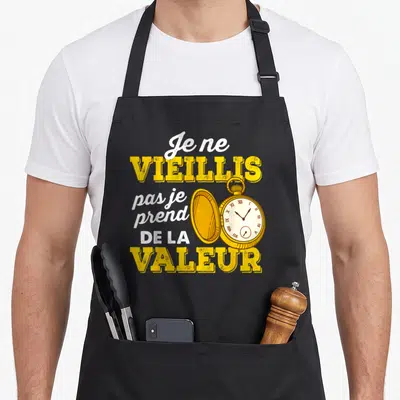 Tablier de cuisine noir avec texte jaune et blanc Je ne vieillis pas je prends de la valeur et une montre à gousset dorée ouverte.