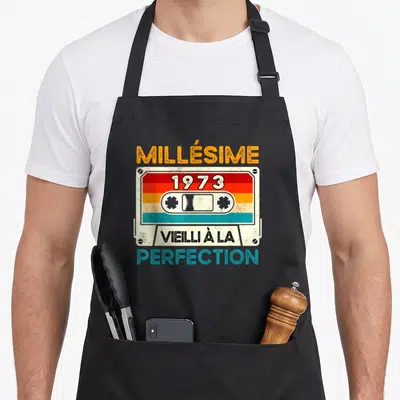 Tablier de cuisine noir avec illustration cassette vintage et texte Millésime 1973 Vieilli à la Perfection en couleurs retro.