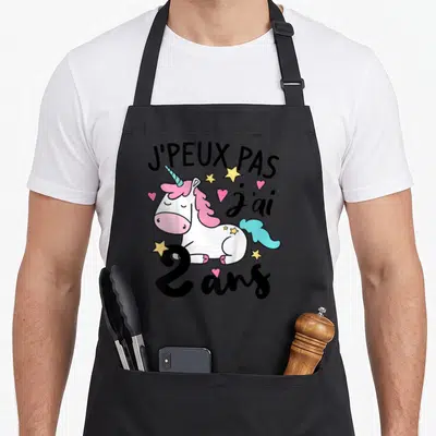 Tablier de cuisine noir avec licorne rose, étoiles jaunes et texte J'peux pas j'ai 2 ans imprimé en noir.