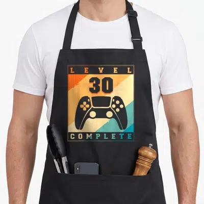Tablier de cuisine noir avec illustration manette de jeu et texte Level 30 Complete sur fond coloré géométrique.