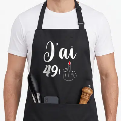 Tablier de cuisine noir avec l'inscription J'ai 49+ accompagnée d'une main au majeur verni en rouge.