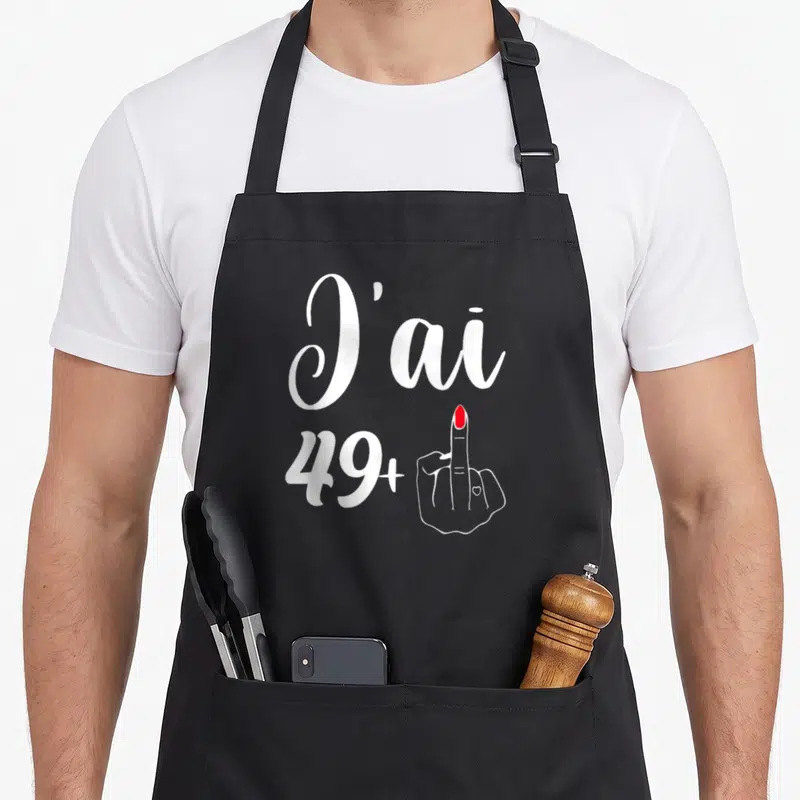 Tablier de cuisine noir avec l'inscription J'ai 49+ accompagnée d'une main au majeur verni en rouge.