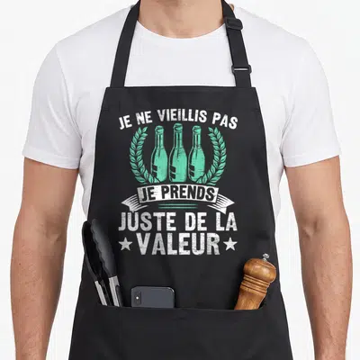 Tablier de cuisine noir avec trois bouteilles vertes encadrées de couronnes et le texte Je ne vieillis pas je prends juste de la valeur en blanc.