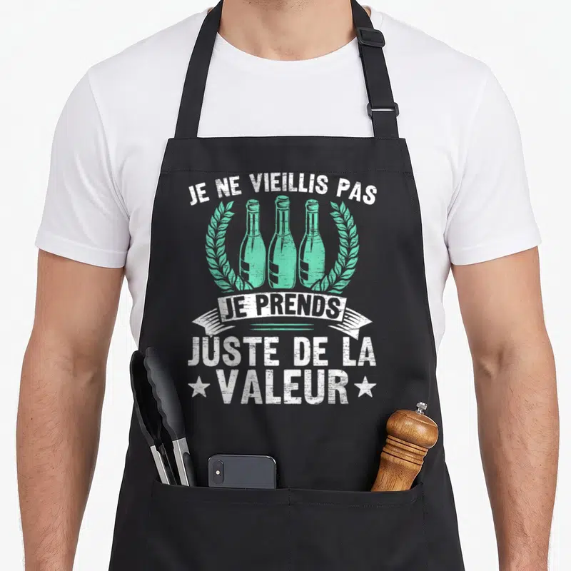 Tablier de cuisine noir avec trois bouteilles vertes encadrées de couronnes et le texte Je ne vieillis pas je prends juste de la valeur en blanc.