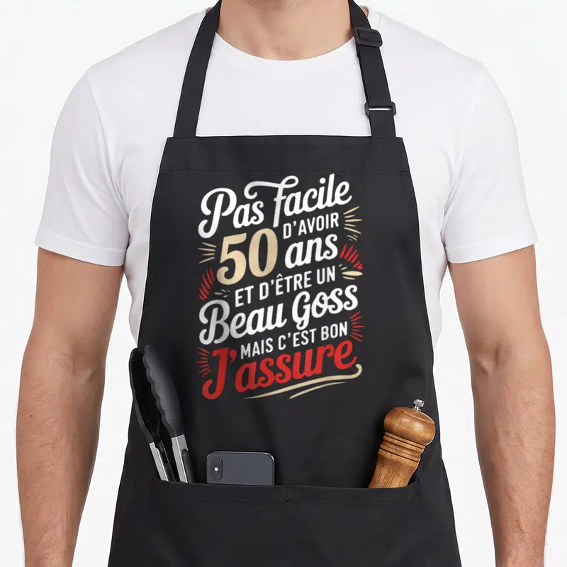 Tablier de cuisine noir avec typographie blanche beige et rouge affichant Pas facile d'avoir 50 ans et d'être un beau goss mais c'est bon j'assure.