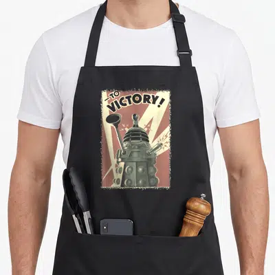 Tablier de cuisine noir avec illustration vintage d'un Dalek et le texte To Victory en arrière-plan rayé rouge.