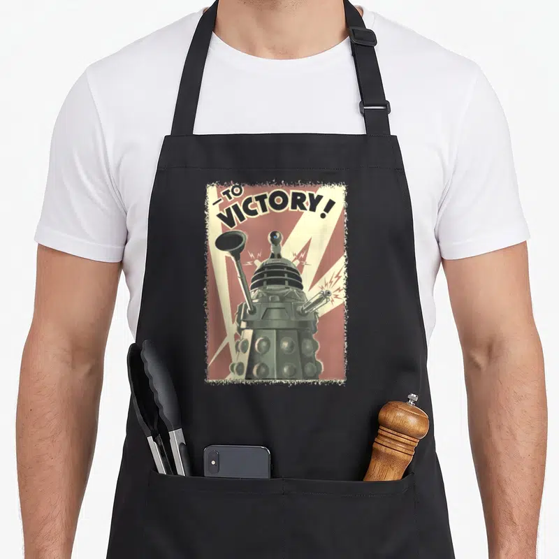 Tablier de cuisine noir avec illustration vintage d'un Dalek et le texte To Victory en arrière-plan rayé rouge.