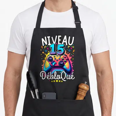 Tablier de cuisine noir avec une manette de jeu colorée et l'inscription Niveau 15 Débloqué en typographie dynamique.