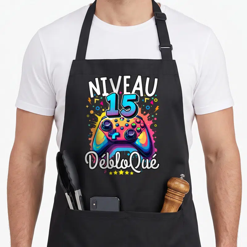 Tablier de cuisine noir avec une manette de jeu colorée et l'inscription Niveau 15 Débloqué en typographie dynamique.