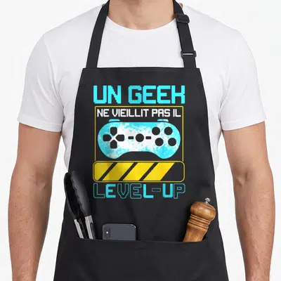 Tablier de cuisine noir avec une manette de jeu vidéo cyan et le texte Un geek ne vieillit pas il level up en lettres pixelisées.