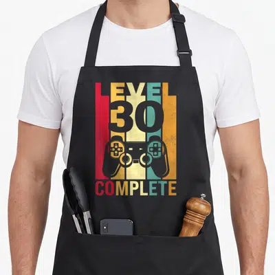 Tablier de cuisine noir avec illustration manette de jeu vidéo et texte Level 30 Complete sur fond coloré vintage.