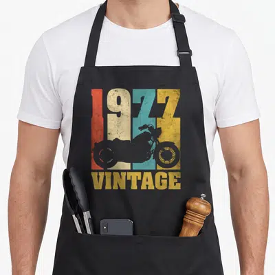 Tablier de cuisine noir avec motif 1977 Vintage Motard Moto aux couleurs délavées et silhouette de moto grise.