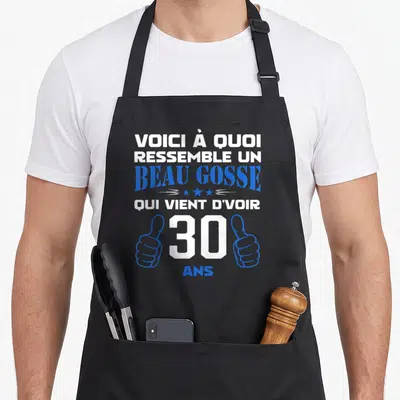 Tablier de cuisine noir avec le texte voici à quoi ressemble un beau gosse 30 ans encadré par deux mains faisant des pouces levés bleus.