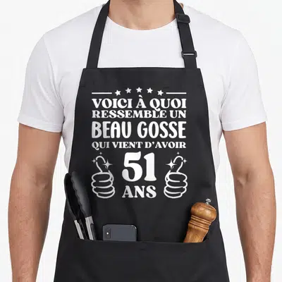 Tablier de cuisine noir avec inscription blanche voici à quoi ressemble un beau gosse qui vient d'avoir 51 ans encadré par deux pouces levés stylisés.
