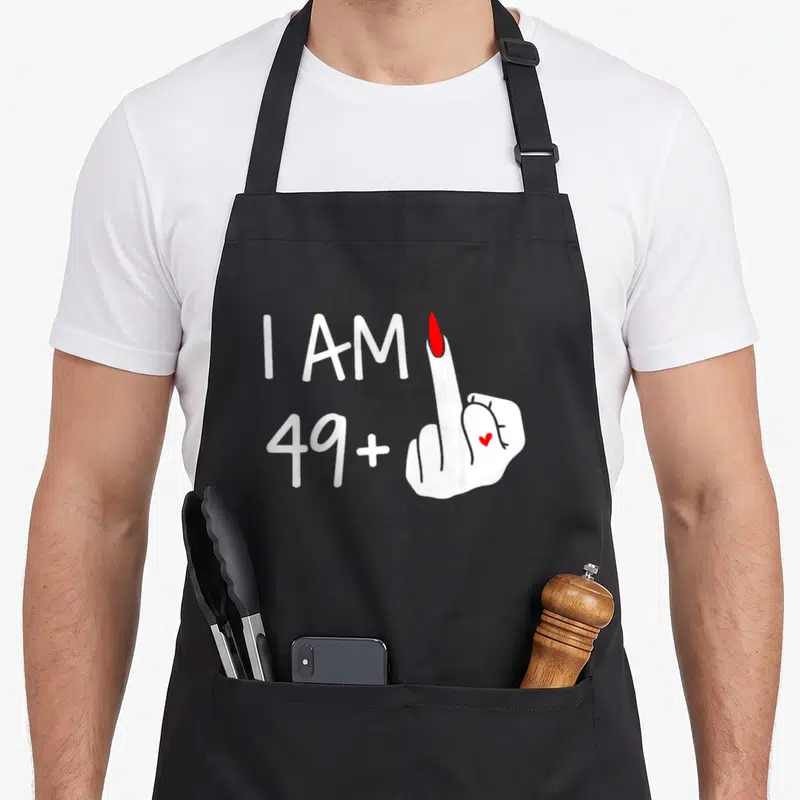 Tablier de cuisine noir avec l'inscription I Am 49 plus un geste d'indifférence et un petit cœur rouge sur la main.