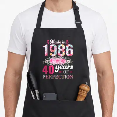 Tablier de cuisine noir avec inscription « Made in 1986 » 40 years of perfection et motifs floraux roses.