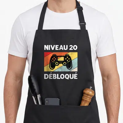 Tablier de cuisine noir avec illustration manette de jeu et texte Niveau 20 Débloqué sur bandes colorées diagonales.