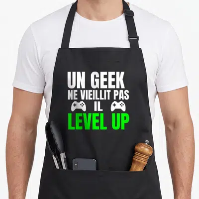 Tablier de cuisine noir avec inscription Un Geek Ne Vieillit Pas Il Level Up et deux manettes blanches.