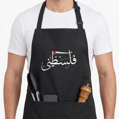 Tablier de cuisine noir avec calligraphie arabe blanche intégrant un petit drapeau palestinien aux couleurs vives.