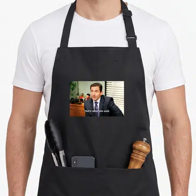 Tablier de cuisine noir avec une image d'un homme en costume et le texte That's what she said imprimé au centre.