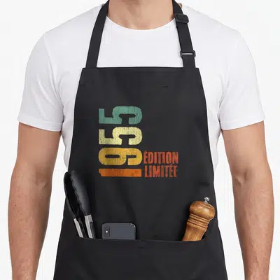 Tablier de cuisine noir avec inscription verticale 70 ans millésime 1955 en couleurs pastel et orange.