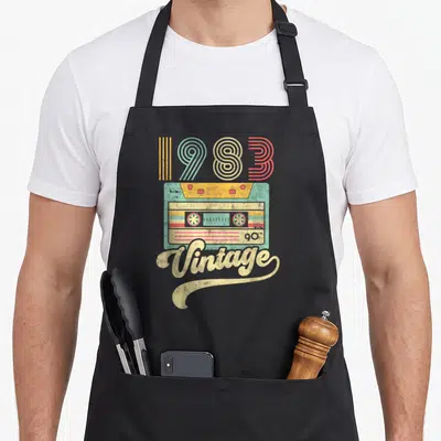 Tablier de cuisine noir avec illustration retro d'une cassette couleur pastel et le texte 1983 Vintage imprimé en grand.