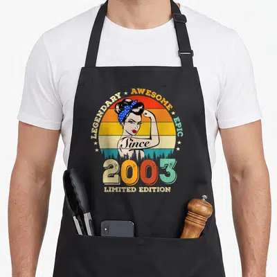 Tablier de cuisine noir avec illustration rétro d'une femme aux cheveux en chignon, bandeau à pois bleus et texteSince 2003 Limited Edition.