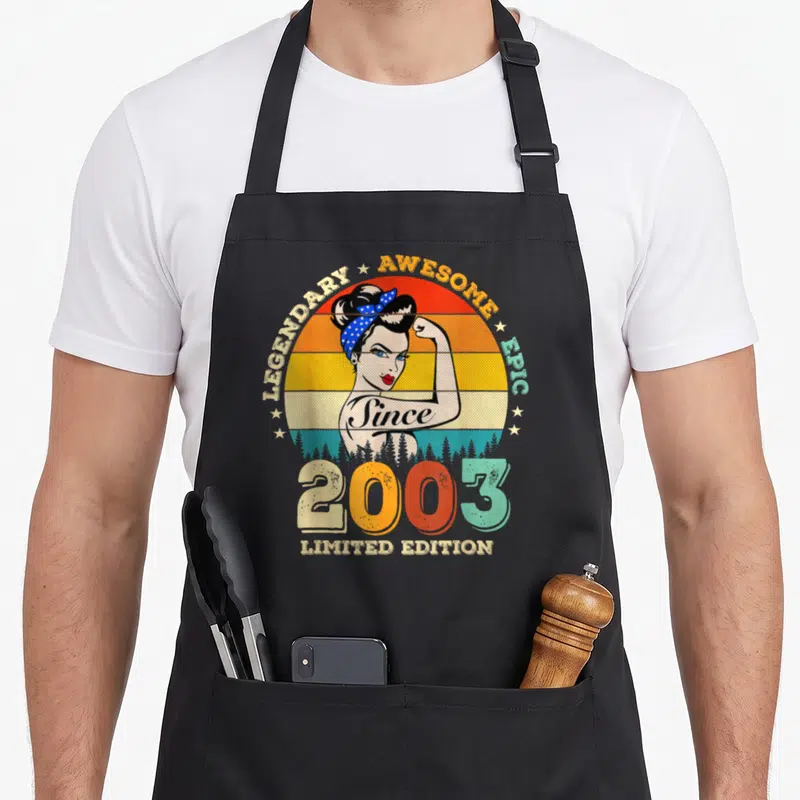 Tablier de cuisine noir avec illustration rétro d'une femme aux cheveux en chignon, bandeau à pois bleus et texteSince 2003 Limited Edition.