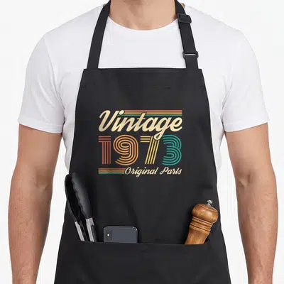 Tablier de cuisine noir avec l'inscription Vintage 1973 Original Parts en typographie rétro multicolore.