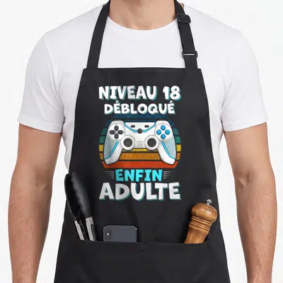 Tablier de cuisine noir avec illustration de manette blanche et texte Niveau 18 débloqué enfin adulte en blanc et bleu cyan.