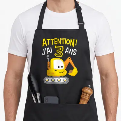 Tablier de cuisine noir avec une pelleteuse jaune souriante et le texte attention j'ai 3 ans imprimé en lettres jaunes et blanches.