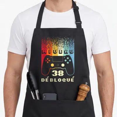 Tablier de cuisine noir avec manette de jeu pixelisée et texte Niveau 38 Débloqué en couleurs vives.