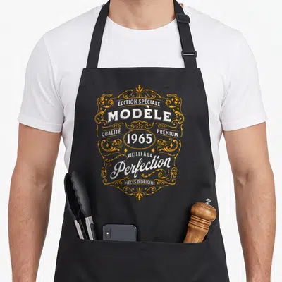 Tablier de cuisine noir avec motif vintage doré et blanc mentionnant Modèle 1965 Vieilli à la Perfection.
