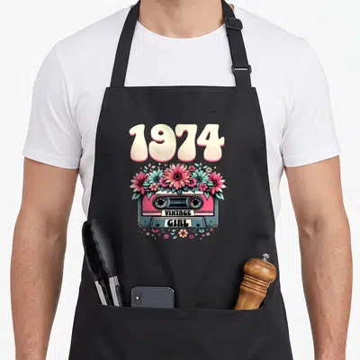Tablier de cuisine noir avec cassette rose, fleurs et l'inscription 1974 vintage girl en lettres arrondies.