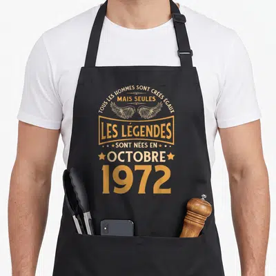 Tablier de cuisine noir avec inscription dorée Les Légendes Sont Nées en Octobre 1972 et illustration d'ailes stylisées au centre.