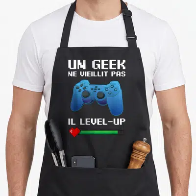 Tablier de cuisine noir avec manette de jeu vidéo bleue, texte blanc pixelisé et barre de vie rouge et verte en bas.