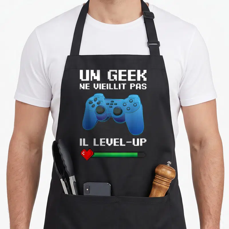 Tablier de cuisine noir avec manette de jeu vidéo bleue, texte blanc pixelisé et barre de vie rouge et verte en bas.