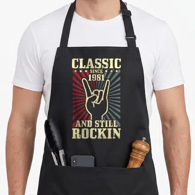 Tablier de cuisine noir avec illustration vintage main rock et texte Classique Since 1981 And Still Rockin en lettres blanches usées.