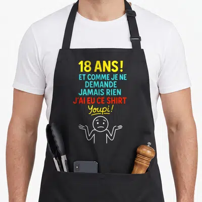 Tablier de cuisine noir avec texte coloré 18 ans et personnage dessiné aux épaules hautes, expression résignée.