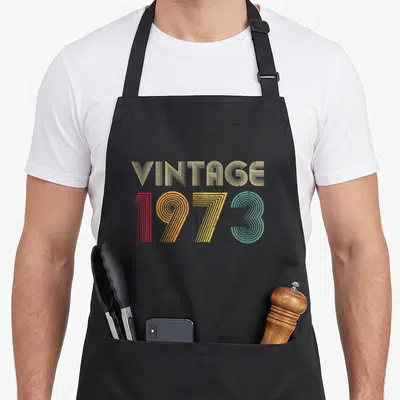 Tablier de cuisine noir avec inscription vintage 1973 aux couleurs rétro et lignes concentriques.