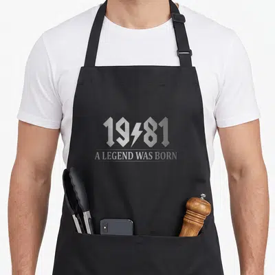 Tablier de cuisine noir avec inscription 1981 une légende was born en typographie argentée stylisée avec un éclair.